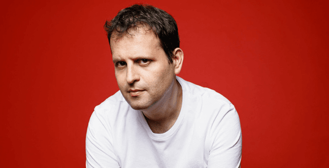 A man with brown hair and a white t-shirt sits in front of a red background looking forward