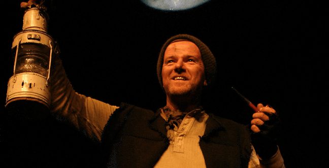 A man holding an oil lamp in the air with one hand and a pipe in the other. He wears a dark gilet and beanie hat with a cream coloured top. The background is black.