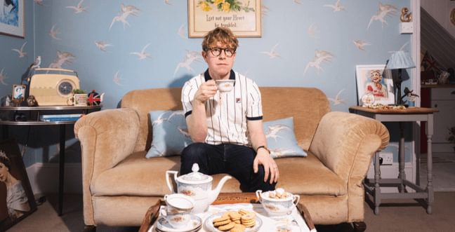 A man with light brown hair and glasses is sitting on a beige sofa in a cozy, vintage-style living room. He is holding a teacup in his right hand and looking straight ahead.