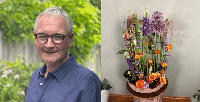 A man with grey hair, wearing black framed glasses and a blue shirt. Next to this is a pot of vibrant flowers with colours of orange, yellow and purple.