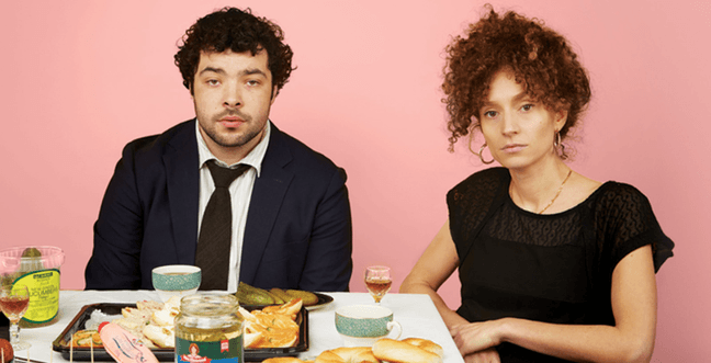 A man in a black suit and tie and a woman in a black dress sit in a pink room at a table of Jewish food