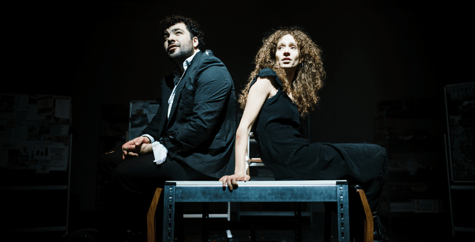 A man in a suit and a woman in a black dress sit back-to-back on a table lit by a single spotlight