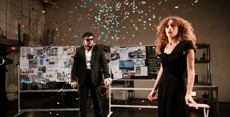 A man in a suit and a woman in a black dress stand in a room surrounded by cork boards as confetti falls around them