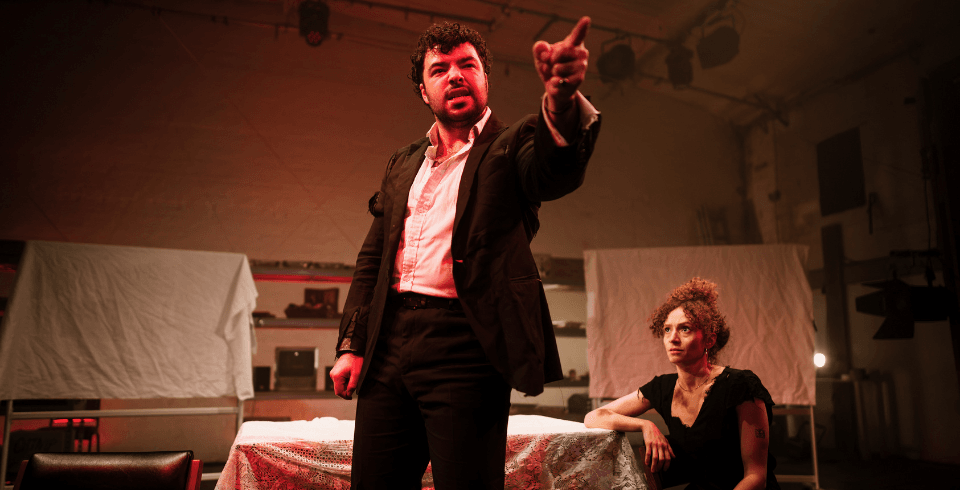 A man in a suit, lit by red light, points out to the audience while a woman crouches behind her