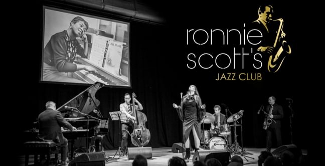 A black and white jazz band on stage with a projection of a singer of a screen above them. The title in the right corner reads 'Ronnie Scott's Jazz Club'
