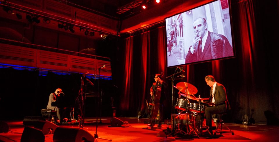 A jazz band on stage in red lights with a black and white photograph projected behind them