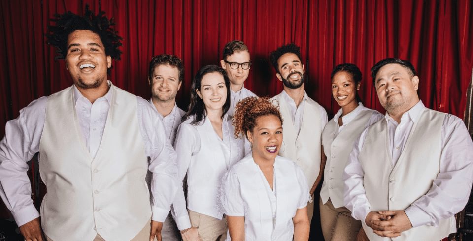 A group of people wearing white shirts stand in front of a red curtain smiling