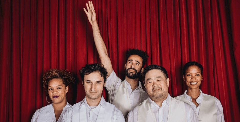 A group of people wearing white shirts stand in front of a red curtain while one man with a dark beard holds his hand in the air