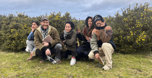 A group of people sat in grass by a large bush and cloudy sky