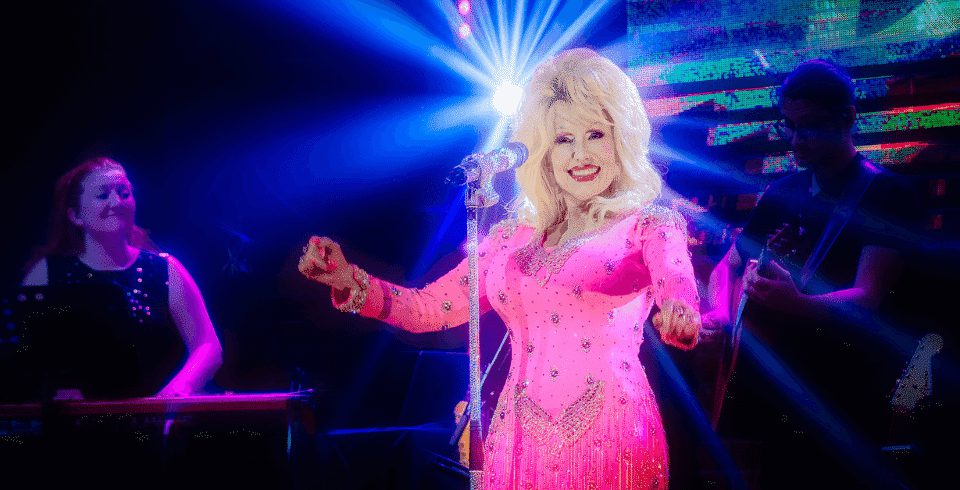 A Dolly Patron tribute with a pink dress, pink nails, and blonde hair stands on stage in front of a microphone.