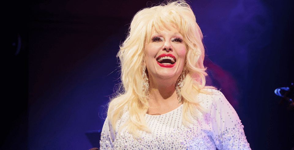 A Dolly Patron tribute in a purple dress on stage, smiles as she looks into the audience.