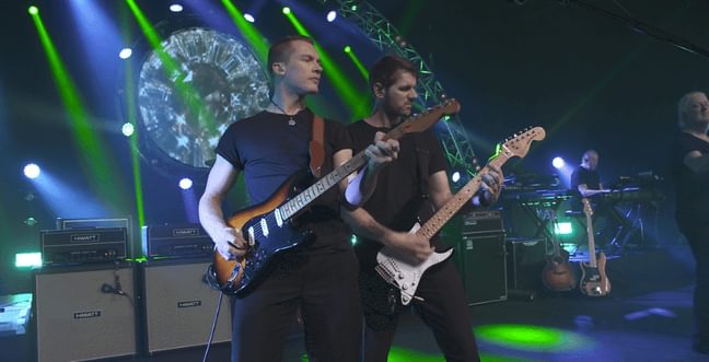 Two guitarists on stage stand side by side in font of a lit-up stage