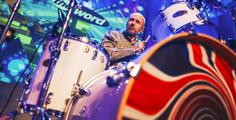 A low angel shot of a drummer performing, the drum kit is white with a swirled Union Jack flag on the front