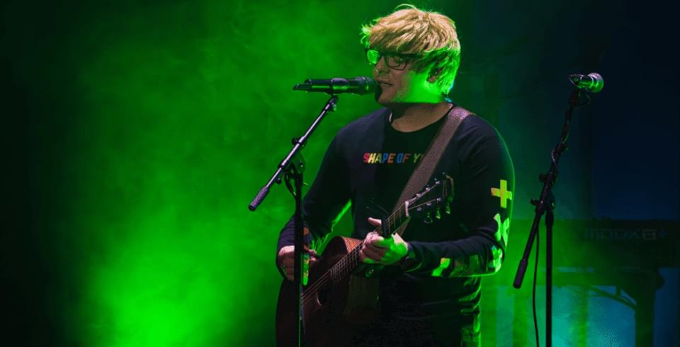 A man with glasses holds a guitar in the green light on stage