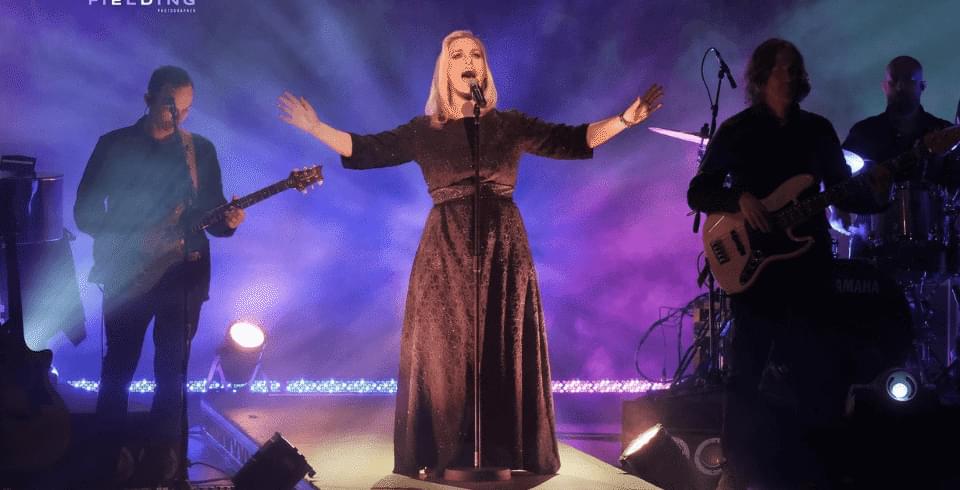A female performer singing into a microphone on stage in a black dress with purple lighting behind her.