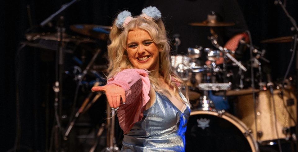 A smiling performer on stage, dressed in a shiny blue outfit with pink sleeves and blue pom-pom hair accessories, extends her hand confidently toward the audience. Drums are visible behind.