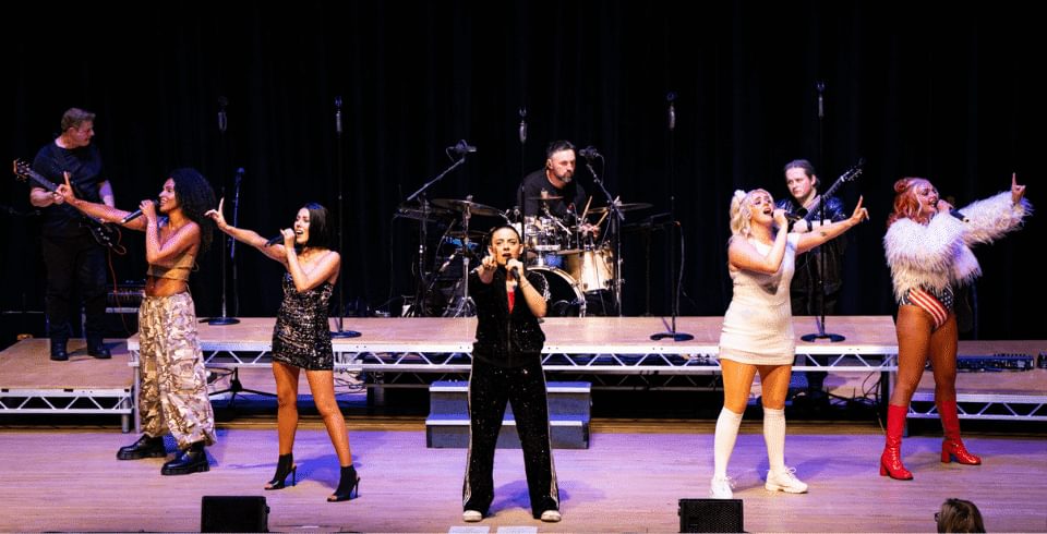 Five performers on stage dressed as spice girls with colourful outfits point in a circle