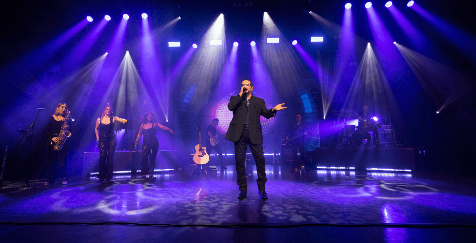 A singer in a black suit and sunglasses performs on stage with a live band, featuring backdrop of blue lights