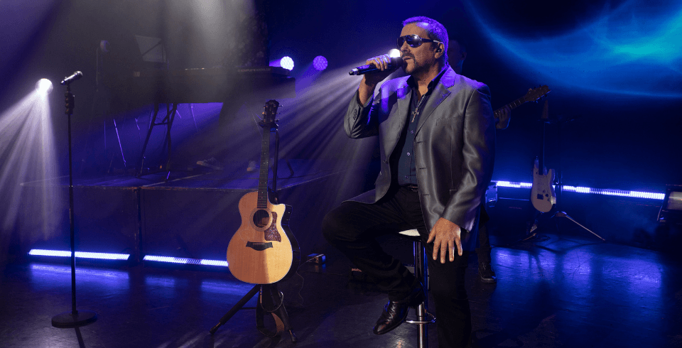 A singer in a black suit and sunglasses performs on stage with a live band, singing into a microphone