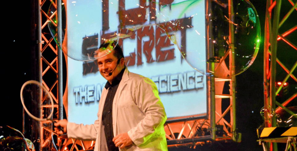A man in a lab coat on stage in front of a sign that says 'Top Secret' and surrounded by large bubbles