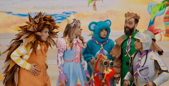 A group of characters in colourful outfits stand together against a painted beach backdrop