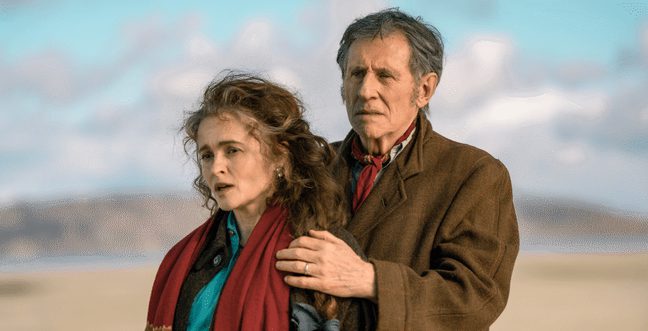A man stood behind a woman, holding her shoulders while they stand on a beach in the daytime.