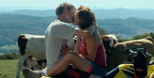 A couple kiss in front of a mountainous backdrop and a field of cows.