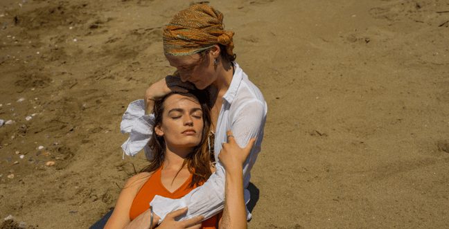 Two women in an embrace, and one leans over the other in the sunshine on a beach