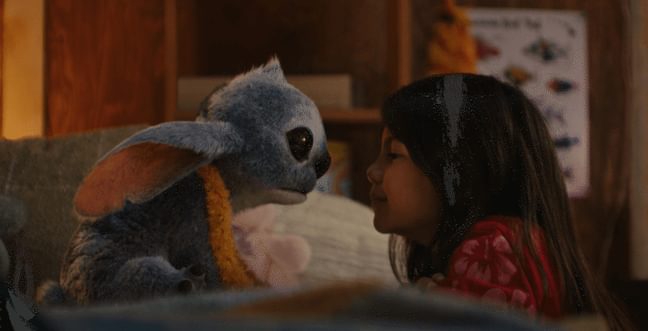A young Hawaiian girl with long brown hair puts her face close to the face of a fluffy blue alien with large rabbit-like ears.