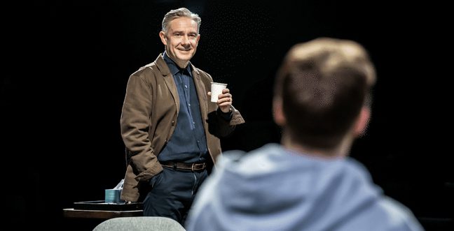 A man wearing a brown jacket and blue shirt smiles while holding a coffee cup, looking towards a man where we can only see the back of his head.