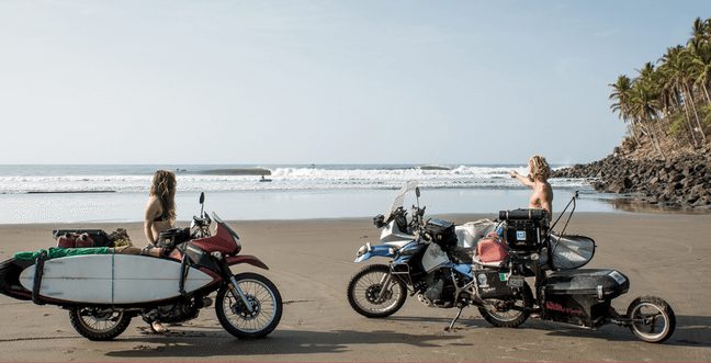 A man and a woman sitting on motorcycles at a beach and looking out to sea.