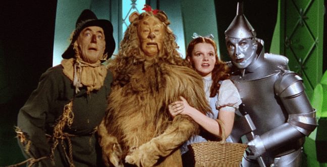 A man dressed as a scarecrow, a man dressed as a lion, a girl in a blue gingham dress and a tinman all stand huddled together.