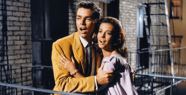 A man in a beige jacket and a woman in a purple dress stand together on a fire escape