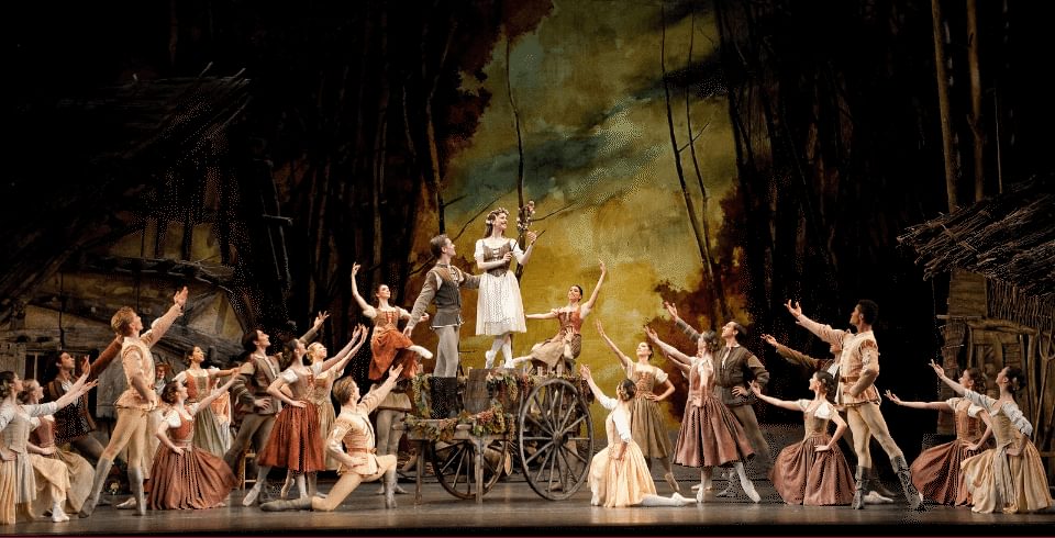 A large group of dancers onstage all pointing towards a female dancer who is standing on a wooden cart.