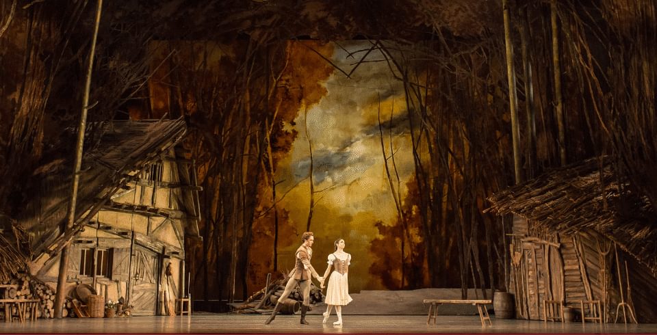 A man and a woman on stage, wearing point-shoes in front of a forest backdrop.