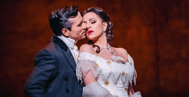 A man wearing a black suit embraces a woman from behind, who is wearing a white dress, standing in front of a red backdrop.