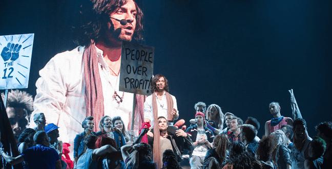 A man in a white top with an orange scarf and long brown hair stands above a crowd of people with a screen of his face behind him