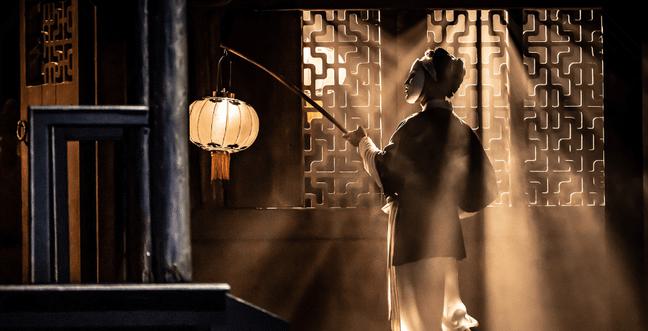 A person holding a warm lamp on a long stick with fringe elements walks into a room with sunbeams through textured window shutters