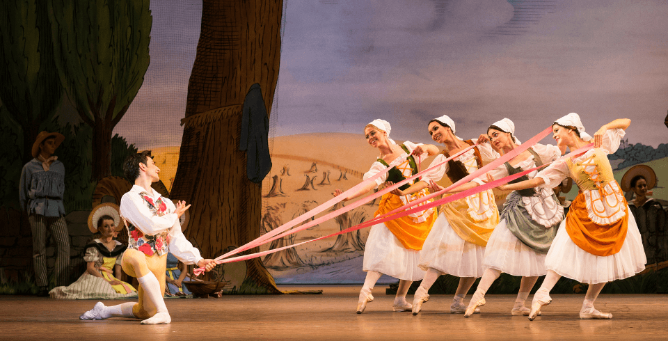 Four women dressed in maid outfits hold onto strands of ribbon, all of which are gathered and held by a man who knees in front of them