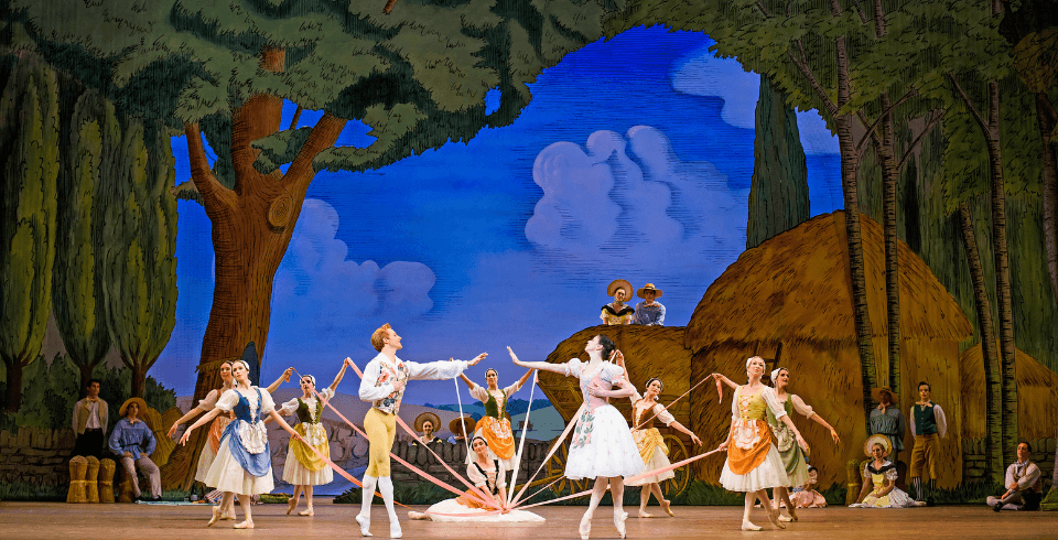 A group of women dressed in maid outfits stand in a circle holding onto strands of ribbon which are gathered together and held towards the ground by a woman sat in the centre of the circle