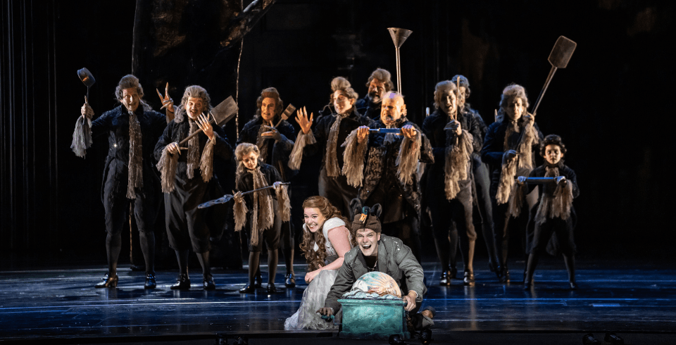 A man and a woman crouch down, smiling at the front of the stage while a group of people in dark clothing all hold various metal tools