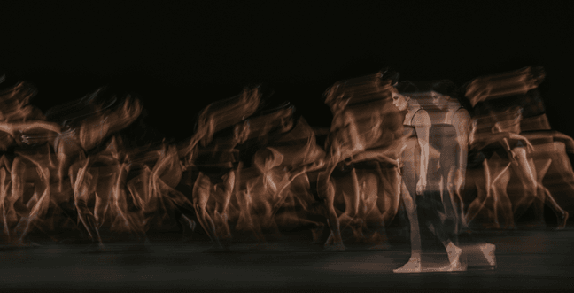 A black background with a group of dancers in motion, captured with a slow shutter speed to create a ghostly, blurred effect that emphasizes movement and fluidity.