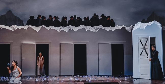 A woman in a white dress holds a purple flower while she sits on stage. She is next to a row of dark doors with a group of people in silhouettes looking down on the stage
