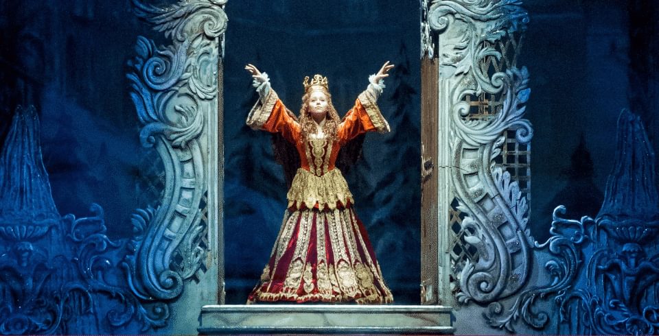 A person in a royal outfit in gold with her arms in the air standing on a large archway of a door