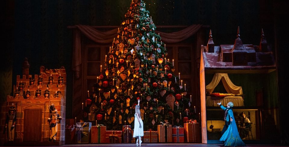 A large Christmas tree on stage with a girl on point shoes in a white dress below
