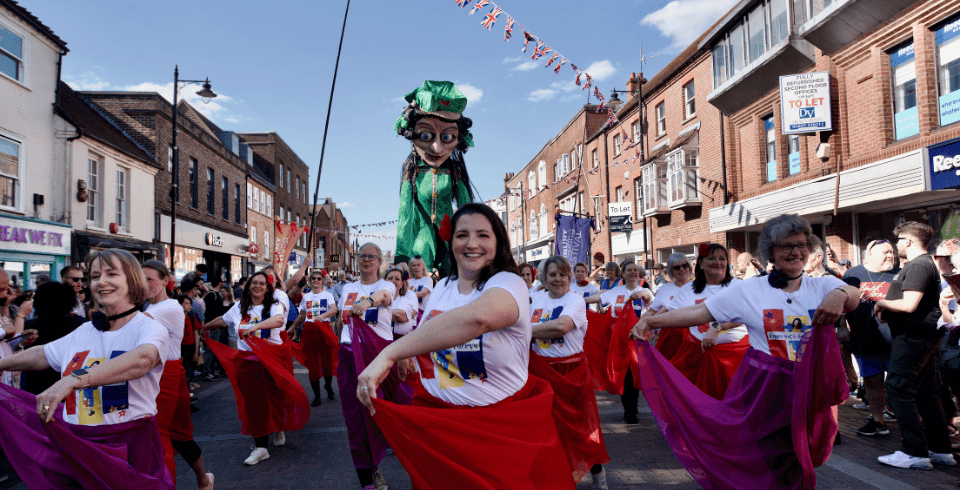 A giant green puppet behind a group of female dancers in colourful skirts.