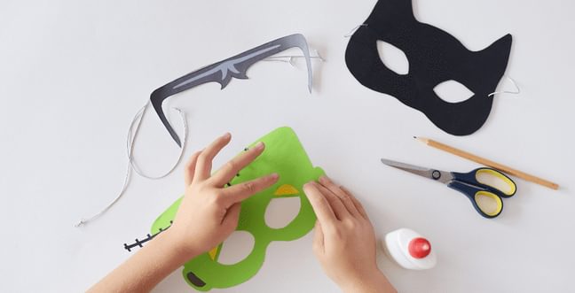 three handmade halloween masks on a white background. Two hands are editing one of them. a pair of scissors, pot of glue and a pencil are also in the shot.