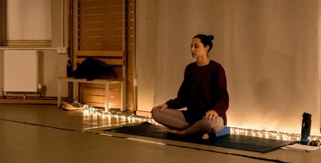 A woman in grey leggings and a dark long-sleeve top sits crossed-legged in front of a string of fairy lights on a mat