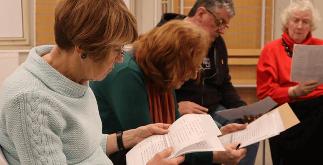 A group of people sit in a circle reading scripts. the woman closest wears a light blue jumper and has short brown hair with glasses
