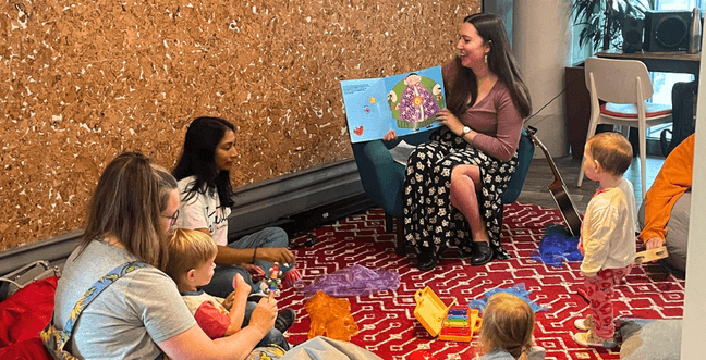 A group of mums and small children sit on a carpet listening to a woman with long brown hair reading a book